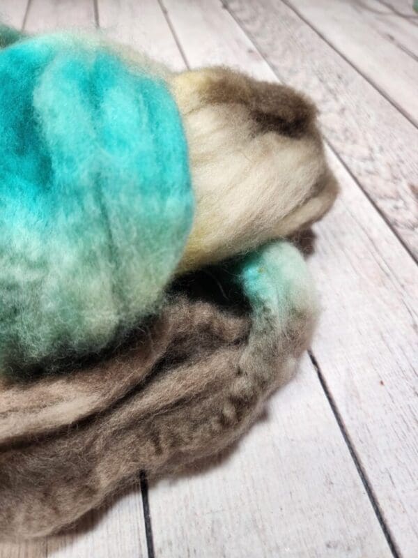 A pile of Disco Date ~ 100g Hand-Dyed Luxury Combed Top Fiber in green, blue, and brown on top of a wooden floor.