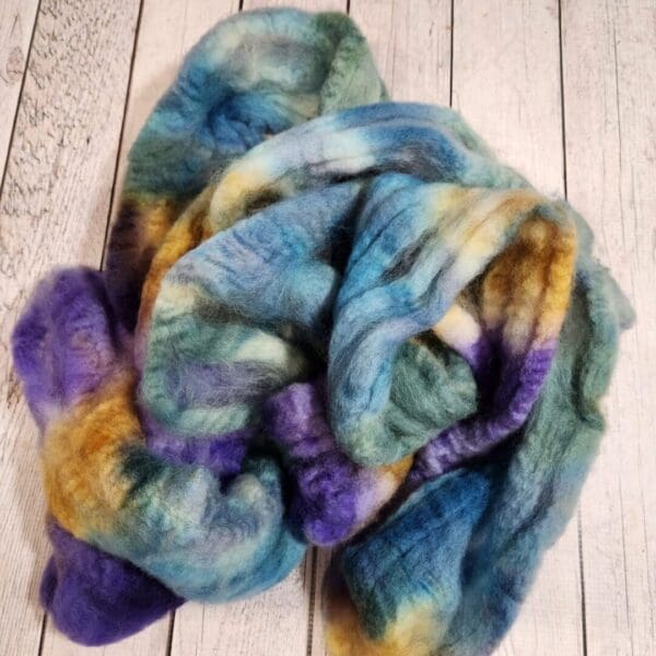 A purple, blue, and yellow Disco Date ~ 100g Hand-Dyed Luxury Combed Top Fiber is laying on a wooden floor.
